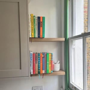 May include: A kitchen scene featuring two wooden shelves holding a collection of cookbooks. The books have colorful spines and titles like "Ottolenghi Simple" and "Jerusalem." A small white mortar and pestle sits on the lower shelf, near a window with green trim.