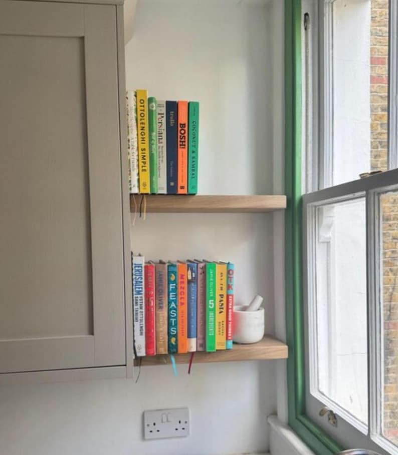 May include: A kitchen scene featuring two wooden shelves holding a collection of cookbooks. The books have colorful spines and titles like "Ottolenghi Simple" and "Jerusalem." A small white mortar and pestle sits on the lower shelf, near a window with green trim.