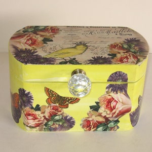 May include: A yellow wooden box with a floral design and a crystal knob. The box has a vintage look and features a bird and text in French.