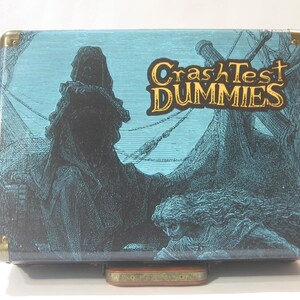 May include: A blue and black suitcase with a graphic of a hooded figure and a ship. The suitcase has gold accents and the text "Crash Test Dummies" in gold lettering.
