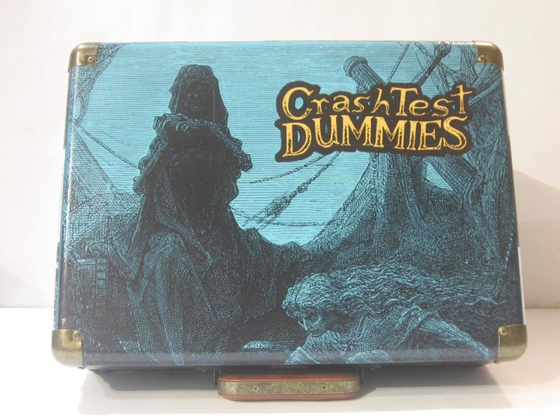 May include: A blue and black suitcase with a graphic of a hooded figure and a ship. The suitcase has gold accents and the text "Crash Test Dummies" in gold lettering.