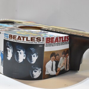 May include: An acoustic guitar with a Beatles album cover design. The guitar is brown with a natural wood finish. The album covers are from the Beatles albums "Meet the Beatles!" and "Help!"