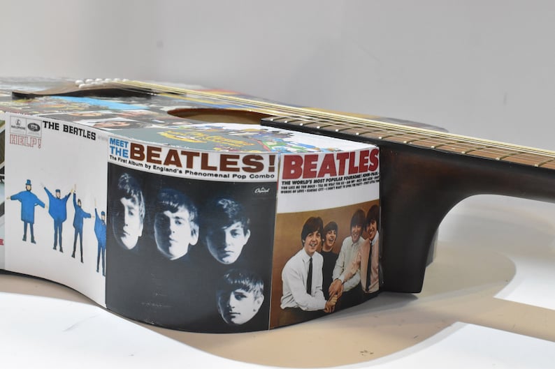 May include: An acoustic guitar with a Beatles album cover design. The guitar is brown with a natural wood finish. The album covers are from the Beatles albums "Meet the Beatles!" and "Help!"
