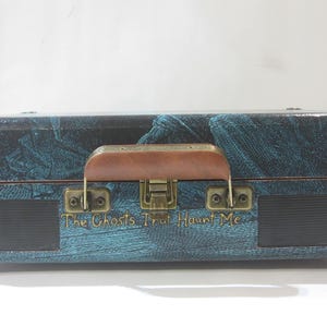 May include: A vintage-style suitcase record player with a teal and black patterned design. The suitcase has a brown leather handle and a brass latch. The text "The Ghosts That Haunt Me" is printed on the front of the suitcase.
