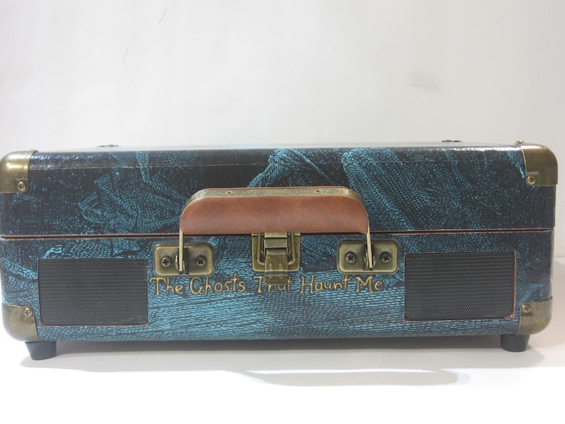 May include: A vintage-style suitcase record player with a teal and black patterned design. The suitcase has a brown leather handle and a brass latch. The text "The Ghosts That Haunt Me" is printed on the front of the suitcase.