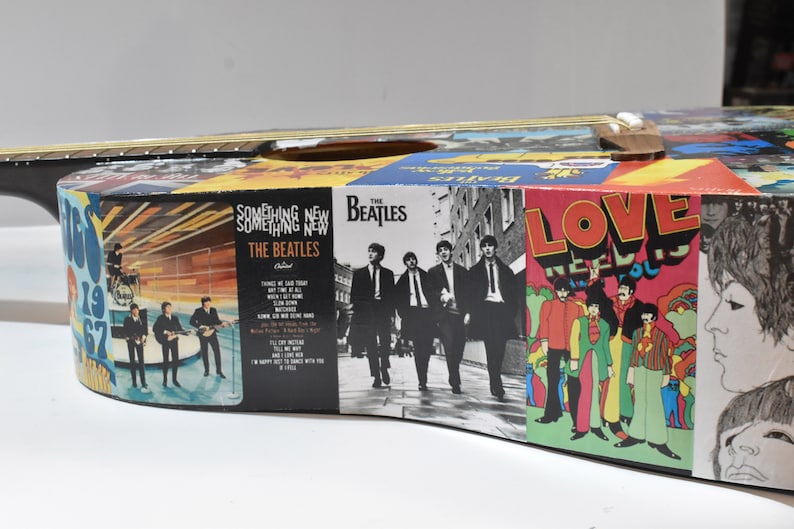 May include: A black acoustic guitar decorated with Beatles album covers. The covers include "Something New" and "Love Needs You".
