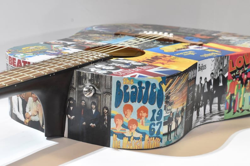 May include: A black acoustic guitar with a colourful collage of The Beatles album covers and images. The guitar has a natural wood finish and a black pickguard.
