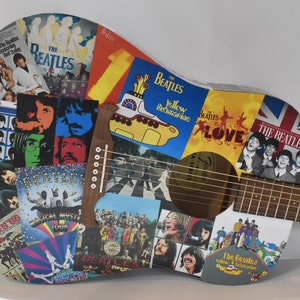 May include: An acoustic guitar decorated with Beatles album covers. The guitar is white with a brown wooden neck and fretboard. The album covers feature images of the band members, album titles, and the band's logo.