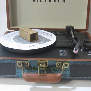 May include: A teal and brown vintage-style record player with a black turntable and a brown handle. The record player has a speaker on the front and the text "The Ghosts Haunt Me" is printed on the front.