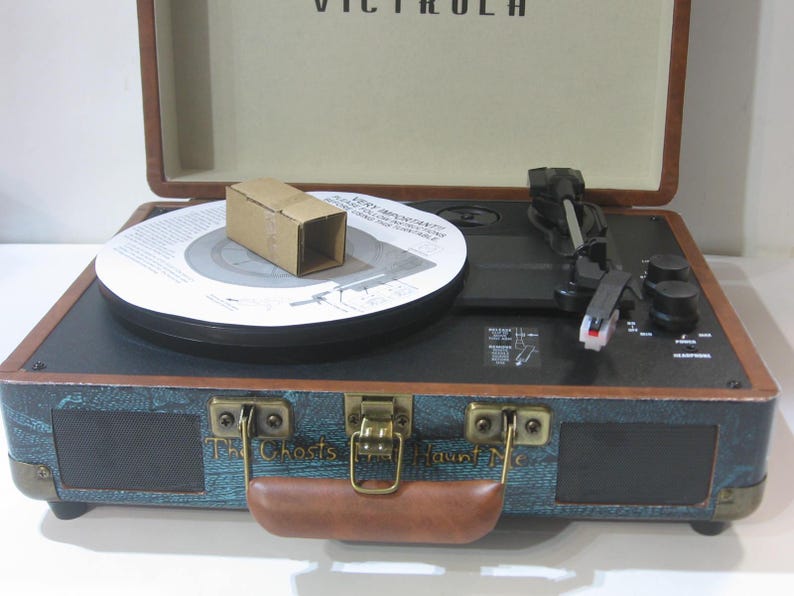 May include: A teal and brown vintage-style record player with a black turntable and a brown handle. The record player has a speaker on the front and the text "The Ghosts Haunt Me" is printed on the front.