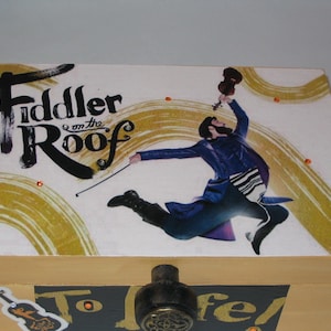 May include: Wooden box with a decorative lid featuring the words "Fiddler on the Roof" and an image of a man playing a violin. The box has a latch and is painted with gold and black accents.