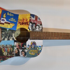 May include: An acoustic guitar with a light blue body covered in Beatles album covers and images. The guitar has a brown wooden neck and a brown wooden headstock with the word "Fender" printed on it.