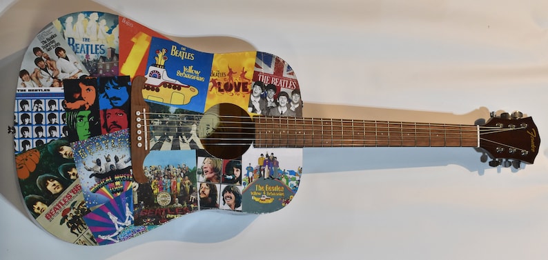 May include: An acoustic guitar with a light blue body covered in Beatles album covers and images. The guitar has a brown wooden neck and a brown wooden headstock with the word "Fender" printed on it.