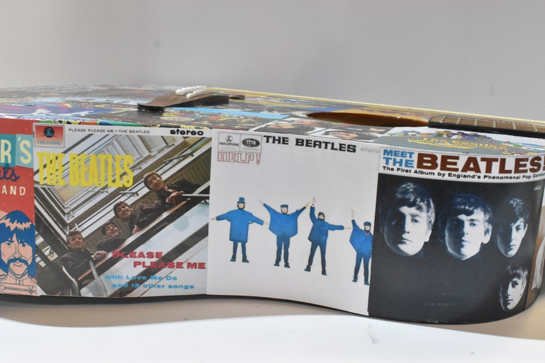 May include: A vintage acoustic guitar decorated with Beatles album covers. The guitar features album art from "Please Please Me", "Help!" and "Meet the Beatles".