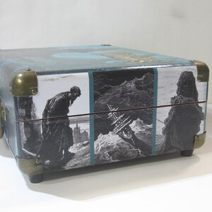 May include: A vintage-style, teal blue, rectangular suitcase with brass hardware and a printed design featuring a man in a long coat, a ship, and a figure in a long robe.