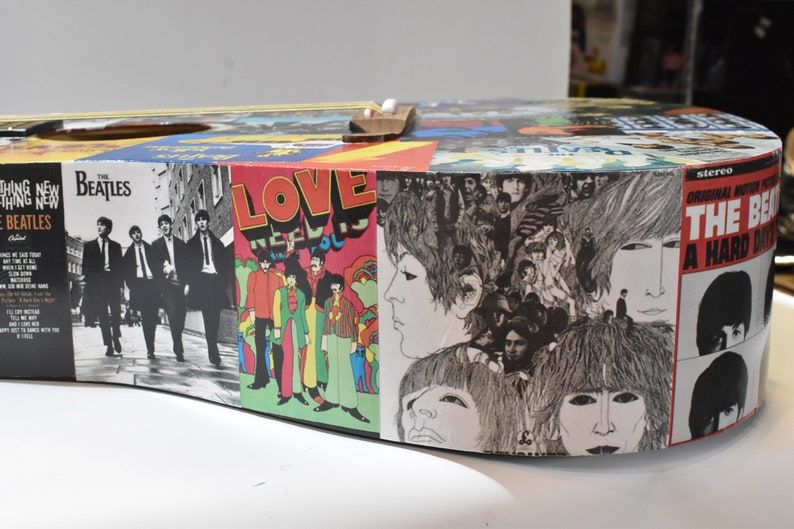 May include: A round, wooden acoustic guitar with a collage of Beatles album covers. The covers include "The Beatles", "Love", "A Hard Day's Night", and "Something New".