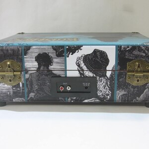 May include: A vintage-style suitcase record player with a blue and black design. The front of the suitcase features a black and white illustration of people. The back of the suitcase has a black panel with audio jacks and a power switch.