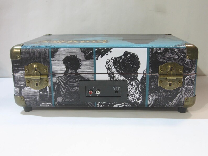 May include: A vintage-style suitcase record player with a blue and black design. The front of the suitcase features a black and white illustration of people. The back of the suitcase has a black panel with audio jacks and a power switch.