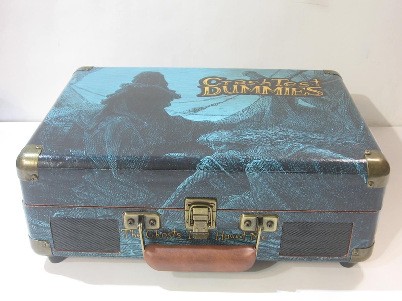 May include: A blue and brown vintage-style record player with a gold handle and metal accents. The top of the record player has a graphic of a ship and the text "Craziest Dummies The Ghosts That Haunt Me".