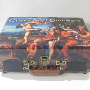 Pode incluir: Um toca-discos de estilo vintage em forma de mala com uma imagem colorida de uma pintura na tampa. A pintura representa um grupo de pessoas em uma paisagem. O texto "Crash Test Dummies" está impresso na tampa em letras douradas.
