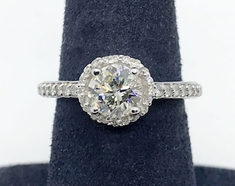 Könnte beinhalten: Ein silberner Verlobungsring mit einem großen runden Diamanten in der Mitte, der von einem Heiligenschein aus kleineren Diamanten umgeben ist. Die Ringschiene ist ebenfalls mit kleineren Diamanten besetzt.