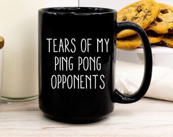 May include: Black ceramic mug with the text "TEARS OF MY PING PONG OPPONENTS" in white. The mug has a large handle and is filled with a dark liquid. Chocolate chip cookies are in the background.