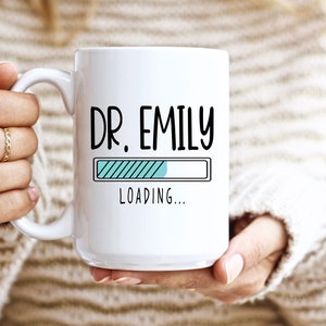 Op de afbeelding: Witte keramische mok met de tekst "DR. EMILY" in het zwart en een laadbalkgrafiek. De mok wordt vastgehouden door een persoon die een trui draagt. De mok is een cadeau voor een dokter.