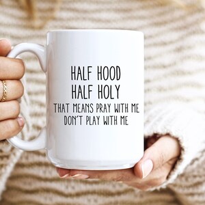 Puede incluir: Taza de cerámica blanca con el texto "HALF HOOD HALF HOLY THAT MEANS PRAY WITH ME DON'T PLAY WITH ME" en negro. La taza la sostiene una persona que lleva un jersey color crema. Anillos dorados adornan los dedos de la persona.
