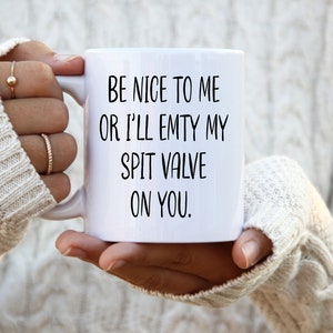 Könnte beinhalten: Weiße Keramik-Tasse mit dem Text "BE NICE TO ME OR I'LL EMPTY MY SPIT VALVE ON YOU" in Schwarz. Die Tasse wird von einer Person gehalten, die einen weißen Zopfstrickpullover trägt.