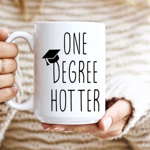 May include: White ceramic mug with the text "ONE DEGREE HOTTER" in black, along with a graduation cap icon. The mug is held by a person wearing a beige sweater.