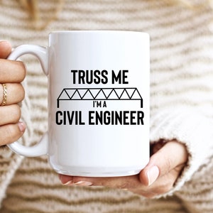 Könnte beinhalten: Weiße Keramiktasse mit dem schwarzen Text "TRUSS ME I'M A CIVIL ENGINEER". Das Design beinhaltet eine Brückengrafik. Die Tasse wird von einer Person gehalten, die einen cremefarbenen Pullover trägt.