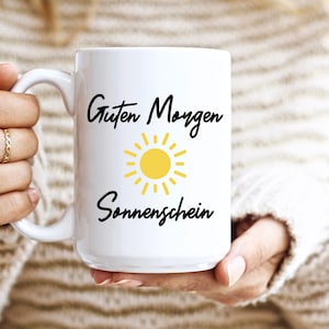 May include: White ceramic mug with the text "Guten Morgen Sonnenschein" and a yellow sun graphic. The mug is held by a person wearing a cream-colored sweater. The mug is a standard size for coffee or tea.