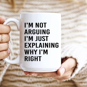 Könnte beinhalten: Weiße Keramik-Tasse mit dem Text "I'M NOT ARGUING I'M JUST EXPLAINING WHY I'M RIGHT" in Schwarz. Die Tasse wird von einer Person gehalten, die einen cremefarbenen Pullover trägt. Die Tasse hat einen Standardgriff.
