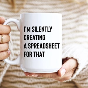 Puede incluir: Una taza de cerámica blanca con el texto "I'M SILENTLY CREATING A SPREADSHEET FOR THAT" en negro. La taza la sostiene una persona que lleva un suéter color crema. La taza es de tamaño estándar.