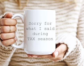 May include: White ceramic mug with the text "Sorry for what I said during TAX season" in black. The mug is held by a person wearing a cream-colored sweater. The mug has a large handle.