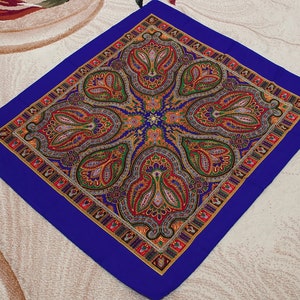 Puede incluir: Un pañuelo cuadrado azul con estampado paisley y borde dorado, rojo y blanco.