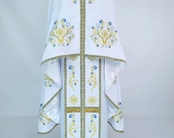 May include: White liturgical vestment with gold and blue embroidered floral designs. The vestment features a high collar, long sleeves, and decorative panels. Gold trim accents the edges and floral patterns. The garment is displayed on a black mannequin head.