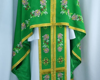 May include: Green liturgical vestments with floral embroidery. The set includes a cape, stole, and undergarment, all adorned with pink and white flowers and gold trim. The fabric is a vibrant green, creating a striking visual contrast.
