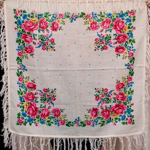 Pode incluir: Um lenço de algodão branco com um padrão floral. O lenço tem uma borda de franjas brancas e um fundo branco com flores rosa, azuis, amarelas e verdes. As flores são dispostas em uma borda ao redor da borda do lenço.