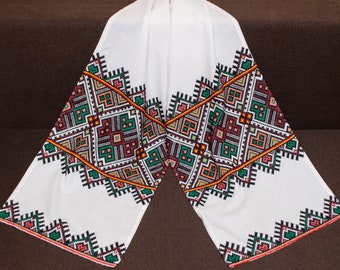 Puede incluir: Tela blanca con un diseño bordado geométrico en negro, verde, naranja, rojo y rosa. El diseño es un patrón repetitivo de cuadrados y triángulos.