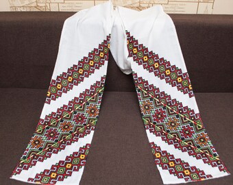 Puede incluir: Tela blanca con patrones geométricos bordados en rojo, verde, amarillo y negro. La tela está doblada por la mitad, con los patrones bordados en ambos lados.
