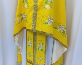 May include: A yellow liturgical vestment with white floral embroidery and gold trim. The vestment has a long, flowing robe with a shorter, fitted under-robe. The fabric is smooth and silky.