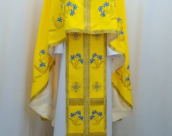 Könnte beinhalten: Eine gelbe Priesterrobe mit blauer Blumenstickerei und goldenen Verzierungen. Die Robe hat ein weißes Untergewand und ein langes, fließendes Design.