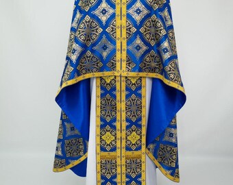 May include: A blue and gold liturgical vestment with intricate patterns. The vestment features a cape-like design with a detailed gold and black brocade pattern over a blue background. The undergarment is white with gold trim.