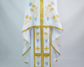 May include: White liturgical vestment with gold and blue embroidery. The vestment features a cape-like top and a long robe, adorned with floral and grape vine patterns. Gold trim edges the garment, adding a touch of elegance.