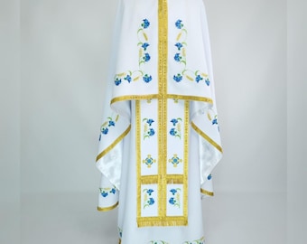 May include: White liturgical vestment with gold trim and blue floral embroidery. The vestment includes a long robe and a cape-like overlay. The gold trim borders the edges and vertical panels, with blue floral patterns throughout.