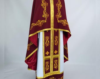 May include: A burgundy liturgical vestment with gold embroidery and trim. The vestment includes a cape, a long white undergarment, and decorative panels. The gold embroidery features floral and scroll designs. The vestment is displayed on a mannequin.