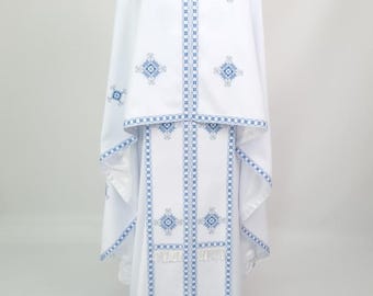 May include: White liturgical vestment with blue embroidered geometric patterns. The vestment includes a long robe, a cape, and a collar, all adorned with intricate designs. The fabric appears to be lightweight and flowing, suitable for religious ceremonies.