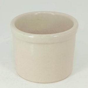 May include: A small, off-white ceramic container with a rounded rim. The cylindrical pot has a smooth, matte finish and a simple, classic design. The interior is visible and appears to be unglazed. This versatile piece could be used for storage or as a decorative item.