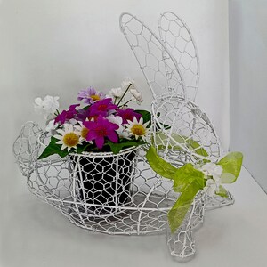 May include: A white wire bunny-shaped planter holding a black pot filled with colorful flowers. The flowers include white daisies, purple, and magenta blooms. A green ribbon and white flowers adorn the bunny.
